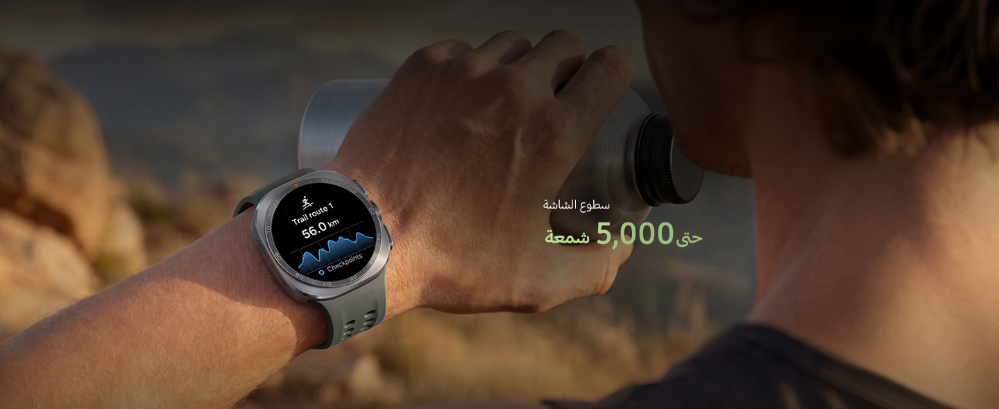 A man on a mountain trail with rocky terrain is holding a water tumbler in his hand while wearing a Galaxy Watch Ultra2 in Titanium Silver with a Marine Band in Olive, displaying a trail route with a distance, ideal for reliable guidance in challenging outdoor conditions during trail running and climbing.