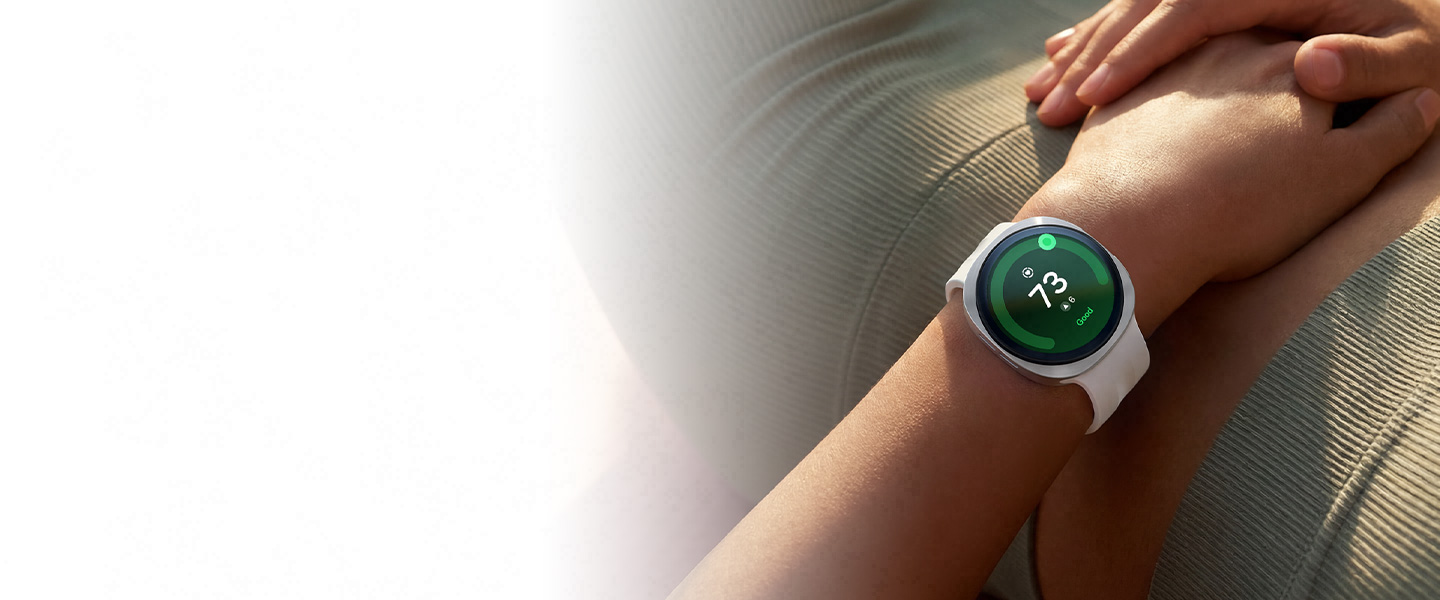 A person lying on their back is wearing Galaxy Watch9 in Cream with a Sport Band in Cream on their wrist and a Heart Health Score of 73 is shown on the watch display, showcasing the heart health tracking capabilities of Galaxy Watch9.